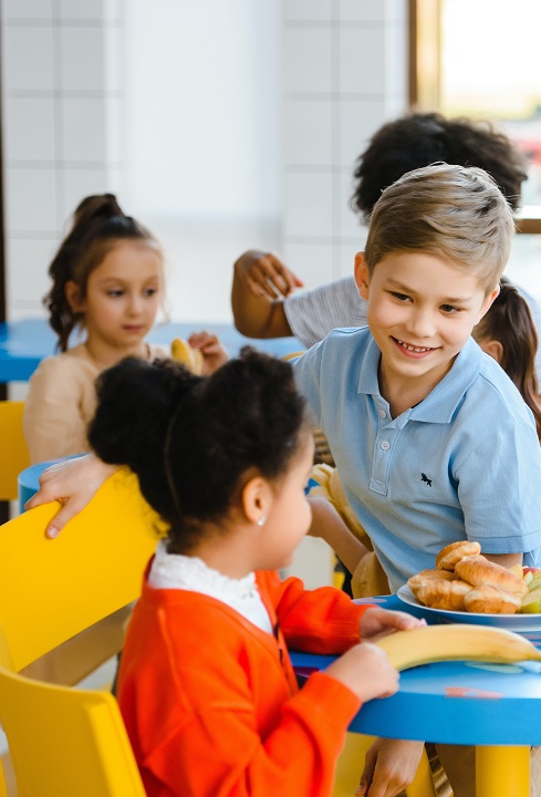 Children sit at a childs dining table in an eating hall which is full of sunlight. The children are touching the food on the table, and talking to each other.