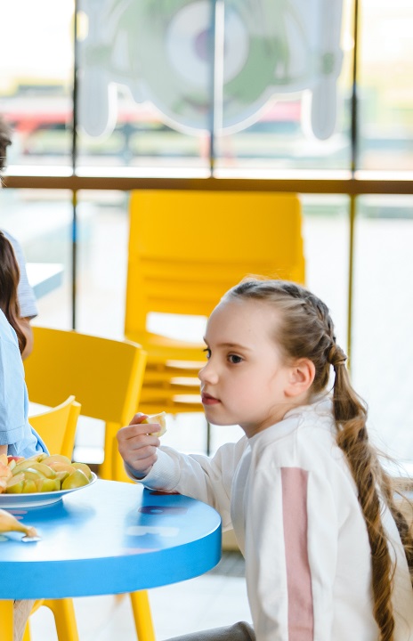 A young girl sits at a childs table in a shared space, holding food in her hand, and staring off into the distance.