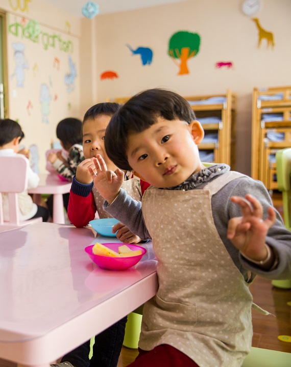 Managing selective eating for autistics: A toddler is sitting at a childrens table and wearing an apron. On the table, the kids are eating freshly cut fruit. The boy is looking at the camera inquisitively.