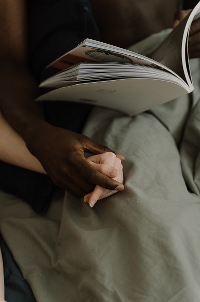 Two adults sit in bed holding hands, one Black and one Caucasian. Only their hands are depicted, with their legs under a gray blanket. The person on the left is reading a magazine. This image was chosen to represent sex and autism sensory sensitivities.